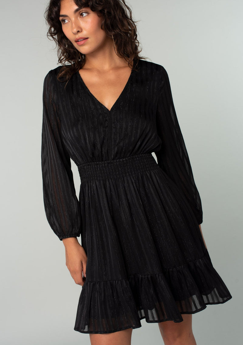 [Color: Black] A front facing image of a brunette model wearing a bohemian holiday mini dress in a black lurex stripe. With long sleeves, a tiered flowy skirt, a smocked elastic waist, a v neckline, and an open back with tie closure.