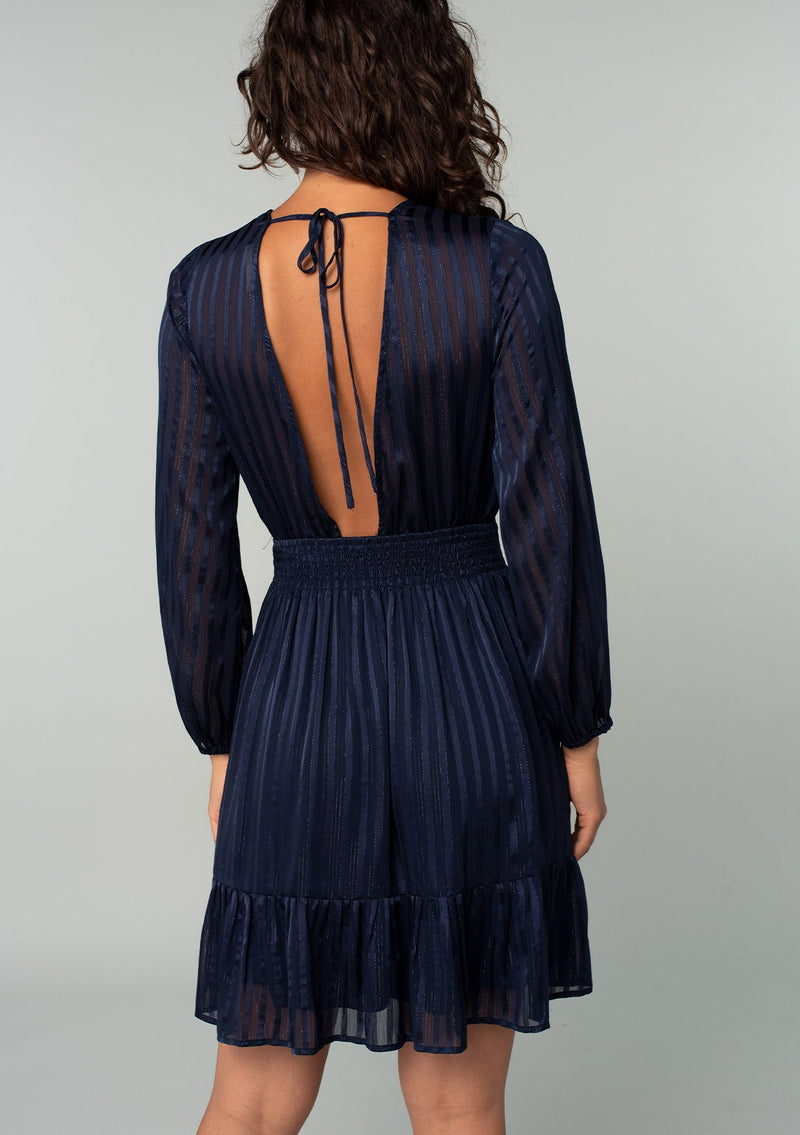 [Color: Navy] A back facing image of a brunette model wearing a bohemian holiday mini dress in a navy blue lurex stripe. With long sleeves, a tiered flowy skirt, a smocked elastic waist, a v neckline, and an open back with tie closure. 