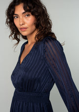 [Color: Navy] A close up side facing image of a brunette model wearing a bohemian holiday mini dress in a navy blue lurex stripe. With long sleeves, a tiered flowy skirt, a smocked elastic waist, a v neckline, and an open back with tie closure. 