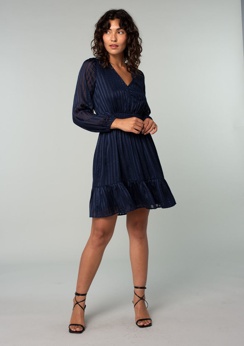 [Color: Navy] A full body front facing image of a brunette model wearing a bohemian holiday mini dress in a navy blue lurex stripe. With long sleeves, a tiered flowy skirt, a smocked elastic waist, a v neckline, and an open back with tie closure.