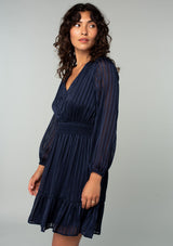 [Color: Navy] A side facing image of a brunette model wearing a bohemian holiday mini dress in a navy blue lurex stripe. With long sleeves, a tiered flowy skirt, a smocked elastic waist, a v neckline, and an open back with tie closure. 