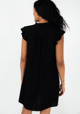 [Color: Black] A back facing image of a brunette model wearing a black bohemian mini dress with a button front, short flutter sleeves, and side pockets.