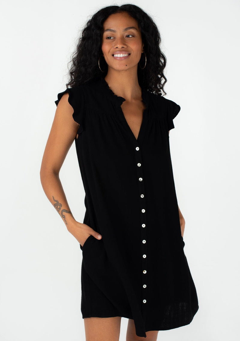 [Color: Black] A half body front facing image of a brunette model wearing a black bohemian mini dress with a button front, short flutter sleeves, and side pockets.