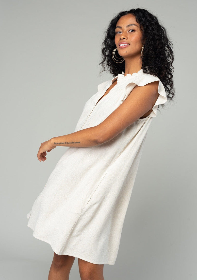 [Color: Natural] A side facing image of a brunette model wearing a natural bohemian mini dress with a button front, short flutter sleeves, and side pockets. 