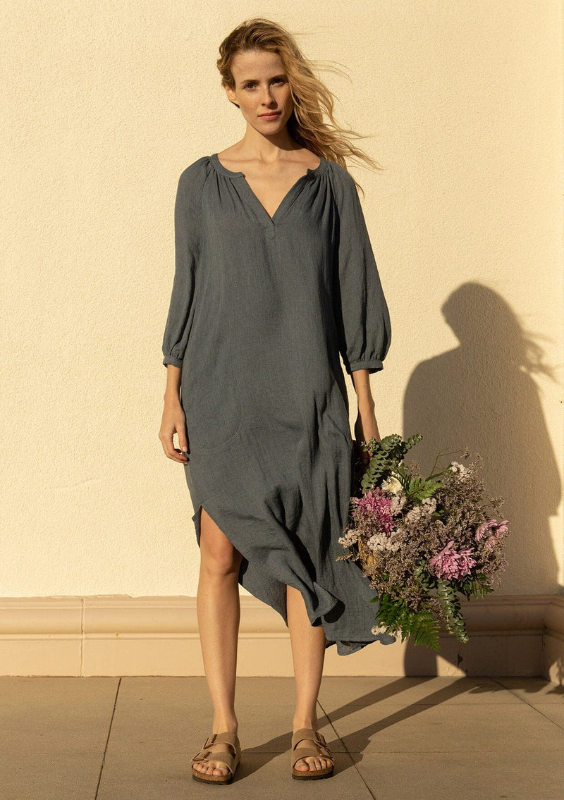 [Color: Navy] A front facing image of a blonde model holding a basket of flowers, wearing a loose, relaxed fit bohemian resort midi dress in navy blue. With three quarter length sleeves, a split v neckline, side pockets, and a loose fit.