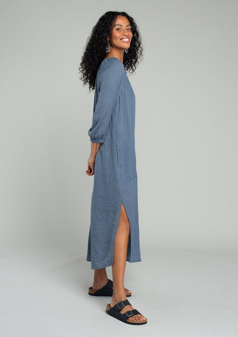 [Color: Navy] A side facing image of a brunette model wearing a loose, relaxed fit bohemian resort midi dress in navy blue. With three quarter length sleeves, a split v neckline, side pockets, and a loose fit. 