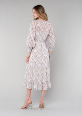 [Color: Ivory/Plum] A back facing image of a blonde model wearing a romantic bohemian mid length chiffon dress in an ivory and plum purple floral print. A spring dress with long sleeves, a self covered button front, a v neckline, a flowy tiered skirt, and a self tie waist belt. 