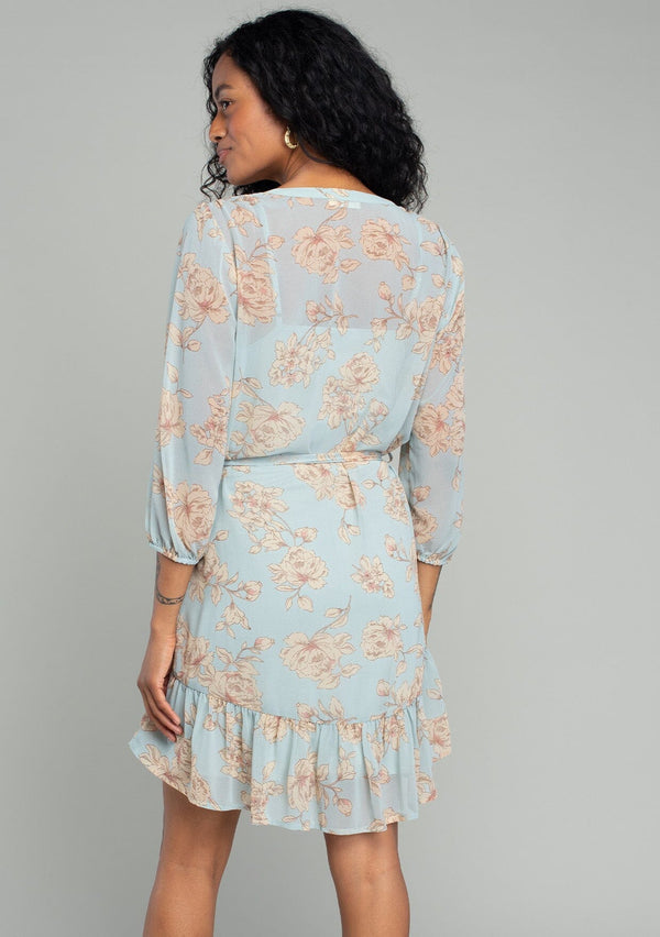 [Color: Dusty Blue/Natural] A back facing image of a brunette model wearing a sheer chiffon bohemian mini dress in a dusty blue and natural floral print. With sheer three quarter length sleeves, a button front, a tiered skirt, and a self tie waist belt. 