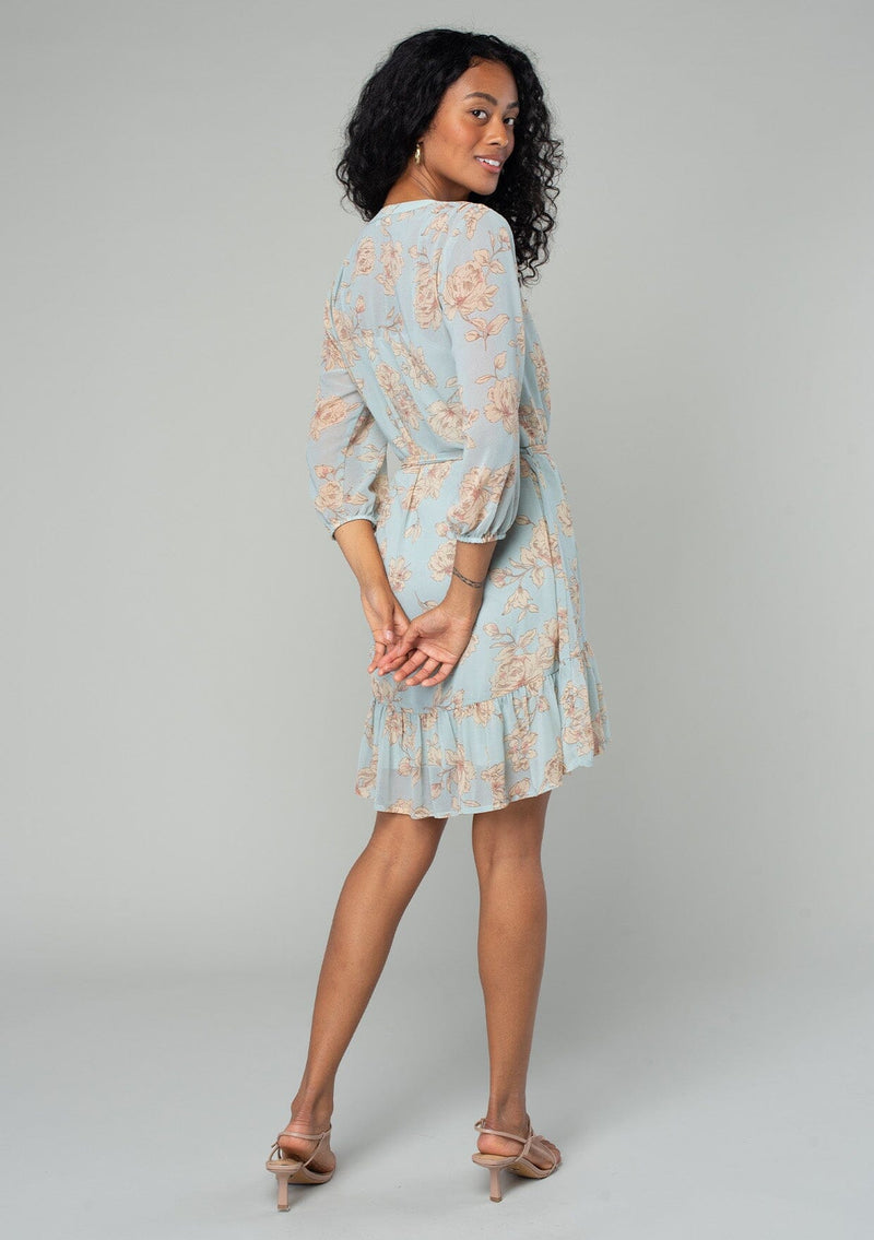 [Color: Dusty Blue/Natural] A back facing image of a brunette model wearing a sheer chiffon bohemian mini dress in a dusty blue and natural floral print. With sheer three quarter length sleeves, a button front, a tiered skirt, and a self tie waist belt. 