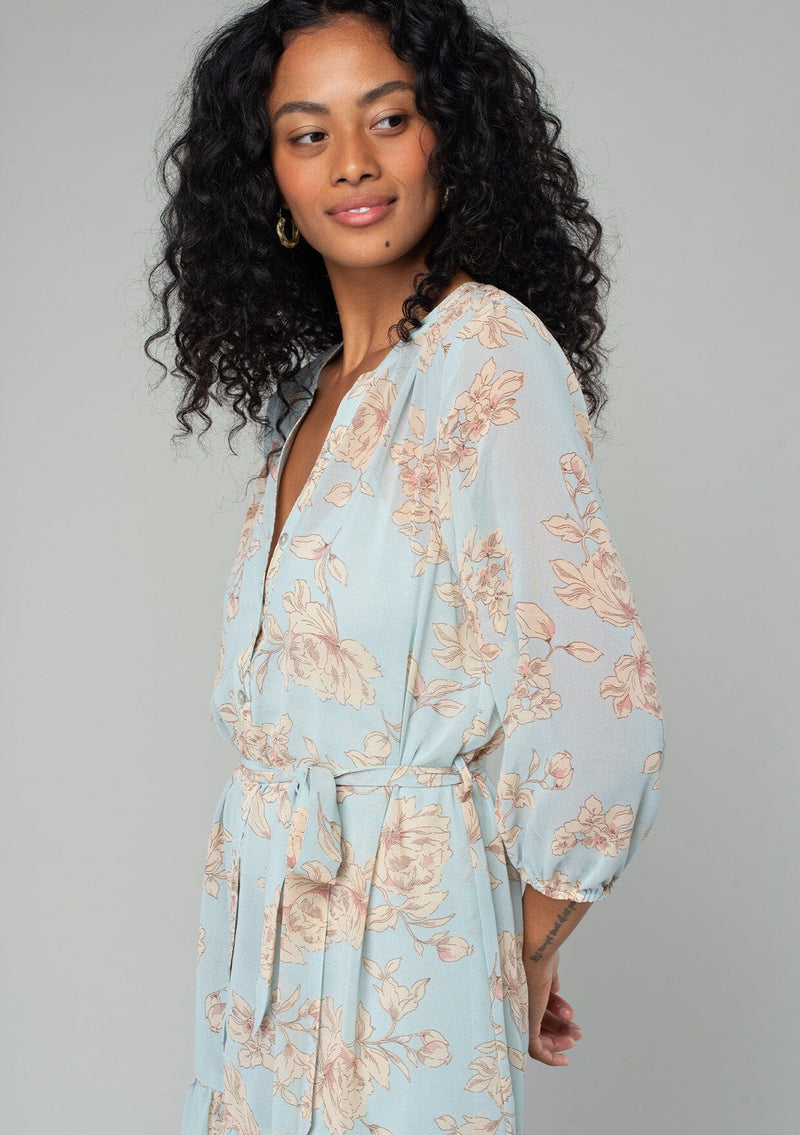 [Color: Dusty Blue/Natural] A close up side facing image of a brunette model wearing a sheer chiffon bohemian mini dress in a dusty blue and natural floral print. With sheer three quarter length sleeves, a button front, a tiered skirt, and a self tie waist belt. 
