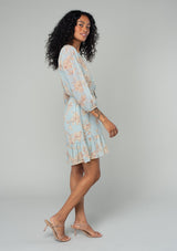 [Color: Dusty Blue/Natural] A side facing image of a brunette model wearing a sheer chiffon bohemian mini dress in a dusty blue and natural floral print. With sheer three quarter length sleeves, a button front, a tiered skirt, and a self tie waist belt. 
