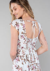 [Color: Mint/Rose] A close up back facing image of a blonde model wearing a spring bohemian mini dress in a mixed pink floral print. With short cap sleeves, a square neckline, an empire waist, a paneled skirt, and a back tie closure. 