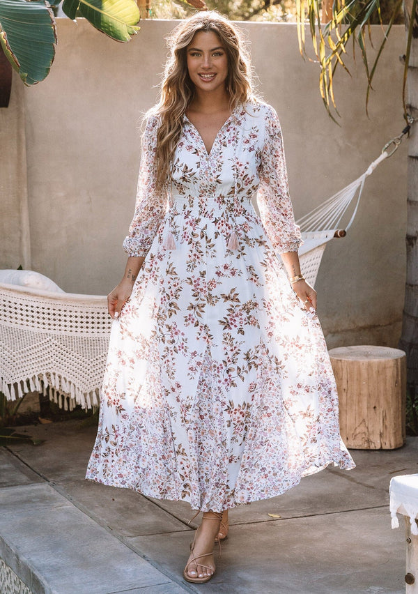 [Color: Mint/Rose] A front facing image of a blonde model outside wearing a classic best selling bohemian maxi dress in a rose pink floral print. A spring maxi dress with long sleeves, a smocked elastic waist, a split v neckline with tassel ties, and a flowy paneled skirt for added shape and movement.
