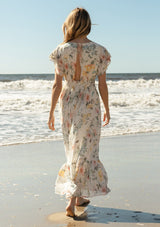 [Color: Natural/Dusty Sage] A back facing image of a blonde model on the beach wearing a romantic chiffon maxi dress in a natural and dusty sage floral print. With short flutter sleeves, a flowy tiered skirt, a back keyhole detail with neck tie closure, and a v neckline. The perfect wedding guest dress.