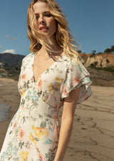[Color: Natural/Dusty Sage] A close up front facing image of a blonde model on the beach wearing a romantic chiffon maxi dress in a natural and dusty sage floral print. With short flutter sleeves, a flowy tiered skirt, a back keyhole detail with neck tie closure, and a v neckline. The perfect wedding guest dress.