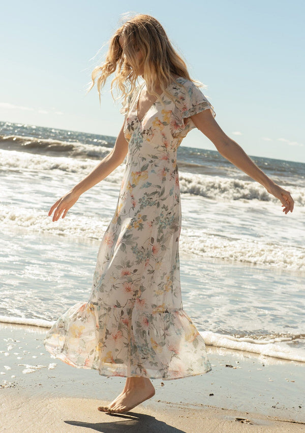 [Color: Natural/Dusty Sage] A side facing image of a blonde model on the beach wearing a romantic chiffon maxi dress in a natural and dusty sage floral print. With short flutter sleeves, a flowy tiered skirt, a back keyhole detail with neck tie closure, and a v neckline. The perfect wedding guest dress.