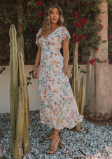 [Color: Natural/Dusty Sage] A side facing image of a blonde model outside wearing a romantic chiffon maxi dress in a natural and dusty sage floral print. With short flutter sleeves, a flowy tiered skirt, a back keyhole detail with neck tie closure, and a v neckline. The perfect wedding guest dress.