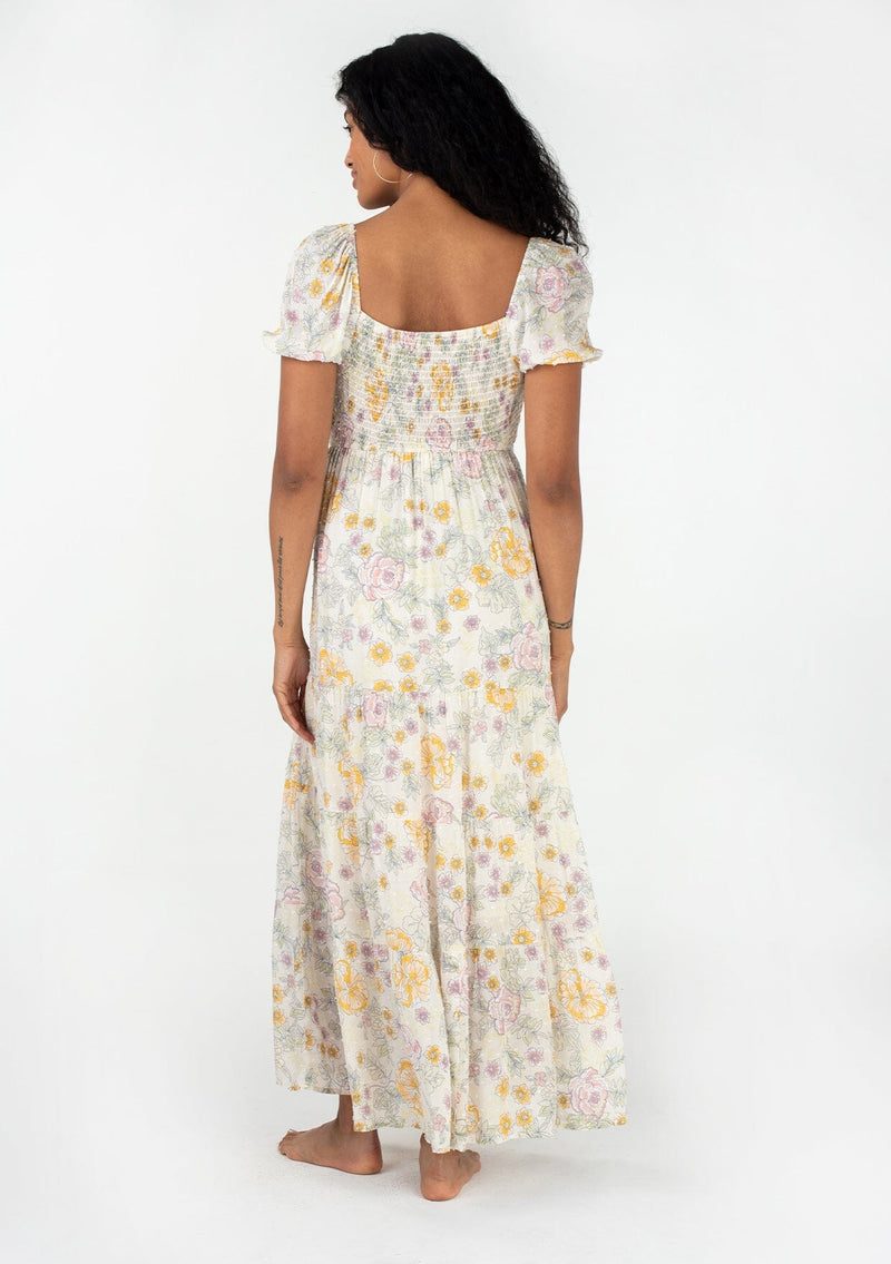[Color: Natural/Pink] A back facing image of a brunette model wearing a bohemian spring maxi dress in an ivory, pink, and yellow vintage inspired floral print. With a sweetheart neckline, short puff sleeves, an empire waist, a flowy tiered skirt, and a smocked elastic bodice at the back. Designed in a pretty textured swiss dot. 
