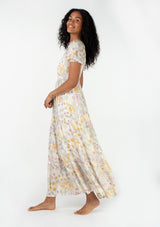 [Color: Natural/Pink] A side facing image of a brunette model wearing a bohemian spring maxi dress in an ivory, pink, and yellow vintage inspired floral print. With a sweetheart neckline, short puff sleeves, an empire waist, a flowy tiered skirt, and a smocked elastic bodice at the back. Designed in a pretty textured swiss dot. 
