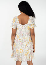 [Color: Natural/Pink] A back facing image of a brunette model wearing a bohemian spring baby doll mini dress in a textured swiss dot. Designed in a vintage inspired floral print, with a flowy silhouette, short sleeves, a square neckline, and a tiered skirt. 