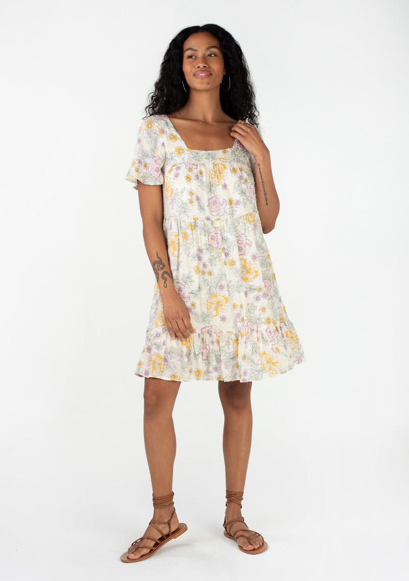 [Color: Natural/Pink] A full body front facing image of a brunette model wearing a bohemian spring baby doll mini dress in a textured swiss dot. Designed in a vintage inspired floral print, with a flowy silhouette, short sleeves, a square neckline, and a tiered skirt. 