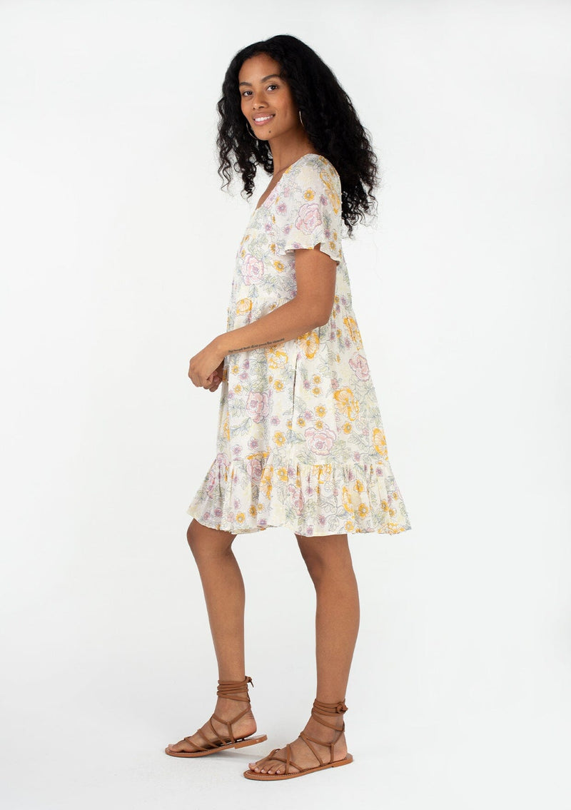 [Color: Natural/Pink] A side facing image of a brunette model wearing a bohemian spring baby doll mini dress in a textured swiss dot. Designed in a vintage inspired floral print, with a flowy silhouette, short sleeves, a square neckline, and a tiered skirt. 