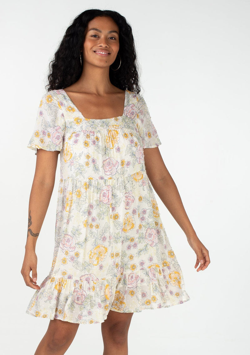 [Color: Natural/Pink] A front facing image of a brunette model wearing a bohemian spring baby doll mini dress in a textured swiss dot. Designed in a vintage inspired floral print, with a flowy silhouette, short sleeves, a square neckline, and a tiered skirt. 