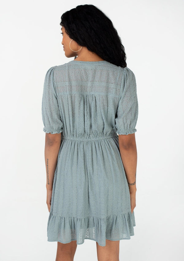 [Color: Dusty Teal] A back facing image of a brunette model wearing a pretty bohemian spring mini dress in a dusty teal eyelet lace chiffon. With short puff sleeves, an elastic ruffled cuff, a button front top, a tiered skirt, and a drawstring waist with tassel ties. 