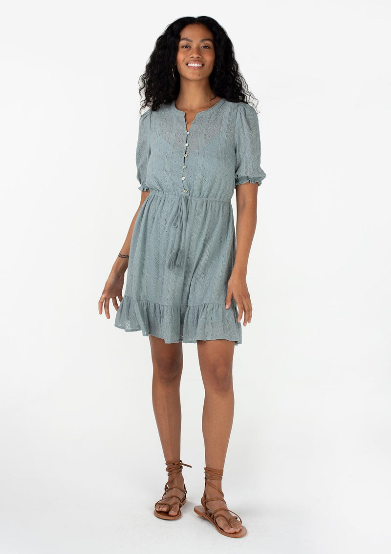 [Color: Dusty Teal] A full body front facing image of a brunette model wearing a pretty bohemian spring mini dress in a dusty teal eyelet lace chiffon. With short puff sleeves, an elastic ruffled cuff, a button front top, a tiered skirt, and a drawstring waist with tassel ties. 