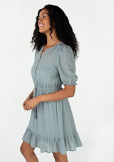 [Color: Dusty Teal] A side facing image of a brunette model wearing a pretty bohemian spring mini dress in a dusty teal eyelet lace chiffon. With short puff sleeves, an elastic ruffled cuff, a button front top, a tiered skirt, and a drawstring waist with tassel ties. 