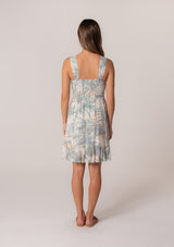 [Color: Natural/Seafoam] A back facing image of a brunette model wearing a sleeveless summer mini dress designed in a green palm leaf print. With a smocked bodice, a square neckline, and a ruffle trimmed tiered skirt. 