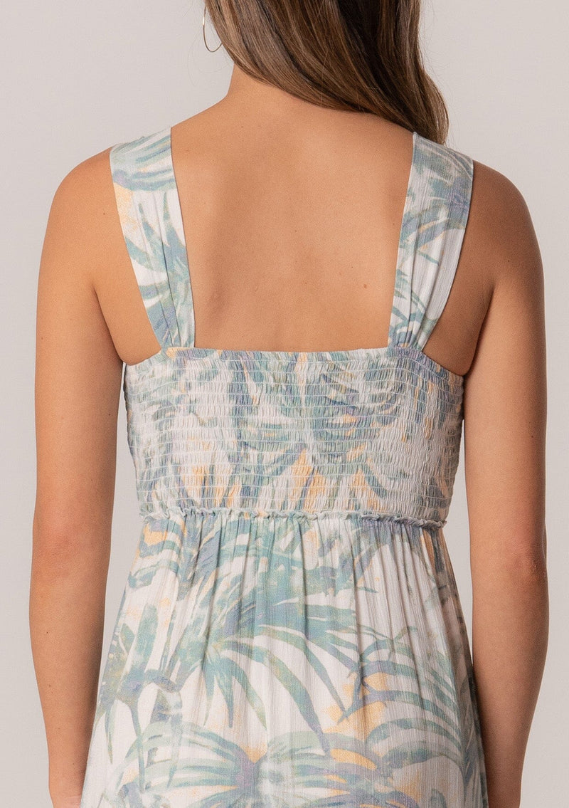 [Color: Natural/Seafoam] A close up back facing image of a brunette model wearing a sleeveless summer mini dress designed in a green palm leaf print. With a smocked bodice, a square neckline, and a ruffle trimmed tiered skirt. 