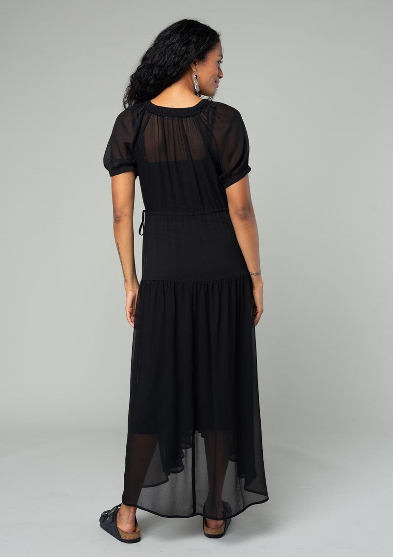 [Color: Black] A back facing image of a brunette model wearing a romantic and flowy black sheer chiffon bohemian maxi dress. With short sleeves, a v neckline, a high low hemline, and a drawstring waist with adjustable side ties. 