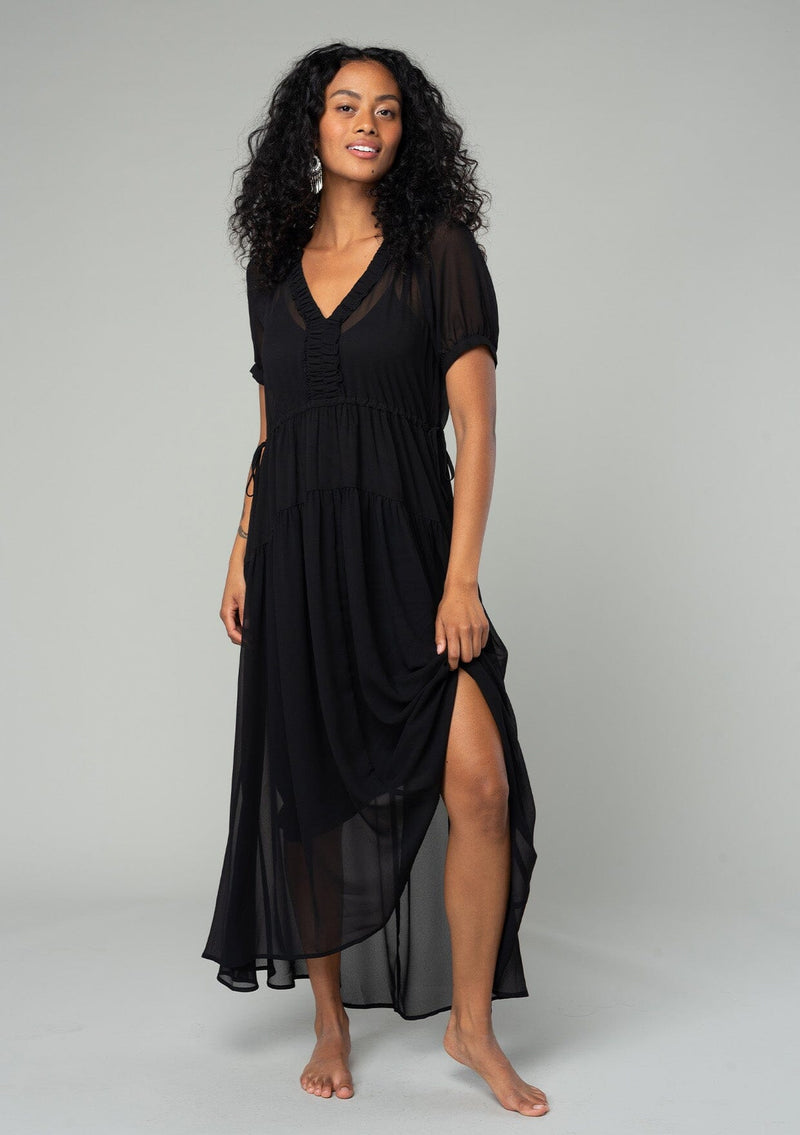 [Color: Black] A full body front facing image of a brunette model wearing a romantic and flowy black sheer chiffon bohemian maxi dress. With short sleeves, a v neckline, a high low hemline, and a drawstring waist with adjustable side ties. 