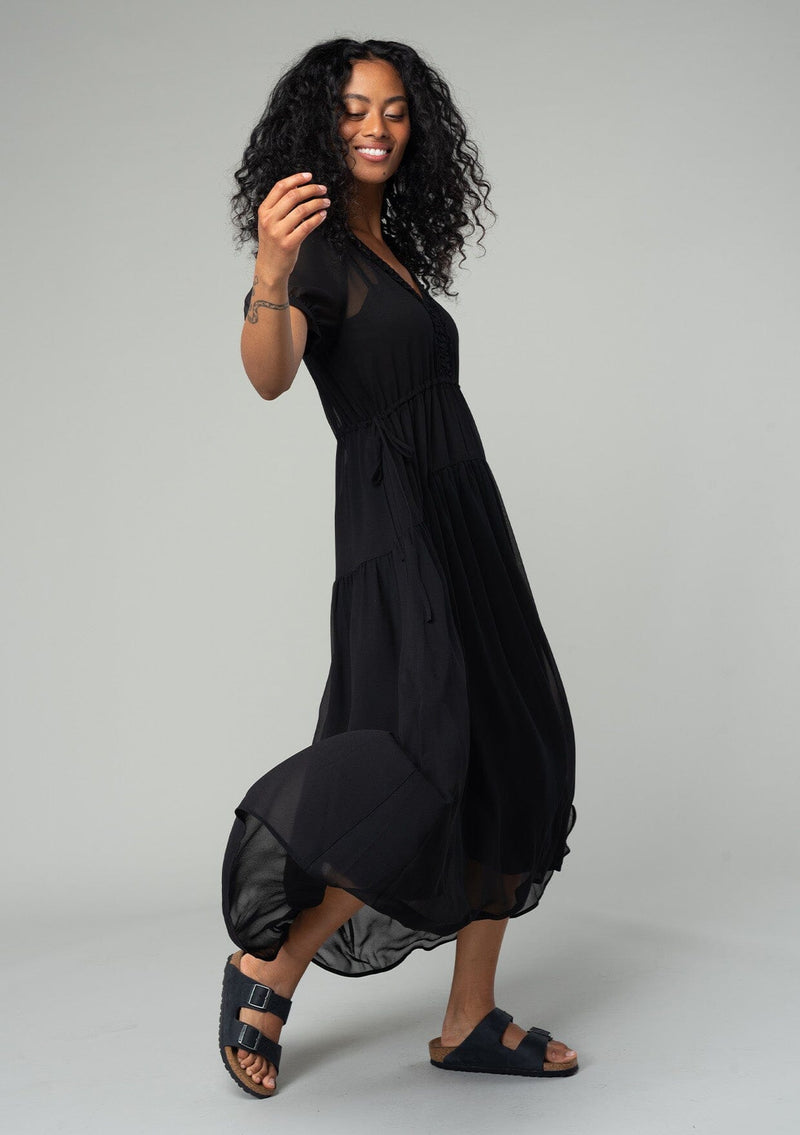 [Color: Black] A side facing image of a brunette model wearing a romantic and flowy black sheer chiffon bohemian maxi dress. With short sleeves, a v neckline, a high low hemline, and a drawstring waist with adjustable side ties. 