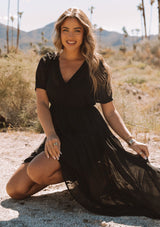 [Color: Black] A front facing image of a blonde model sitting outside wearing a romantic and flowy black sheer chiffon bohemian maxi dress. With short sleeves, a v neckline, a high low hemline, and a drawstring waist with adjustable side ties.