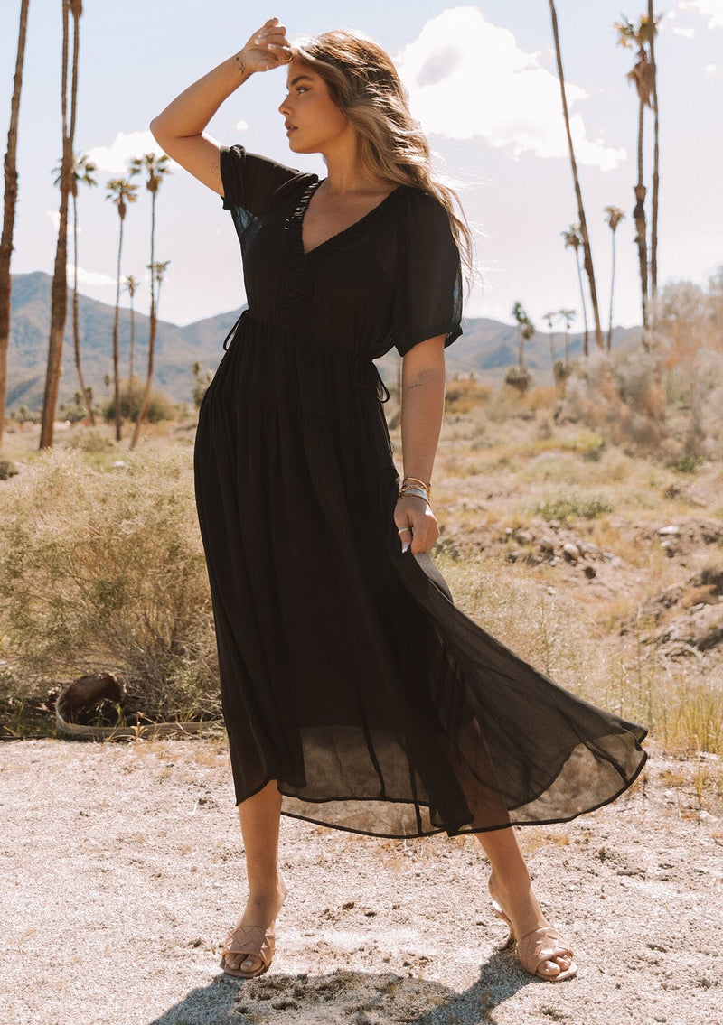[Color: Black] A full body front facing image of a blonde model outside wearing a romantic and flowy black sheer chiffon bohemian maxi dress. With short sleeves, a v neckline, a high low hemline, and a drawstring waist with adjustable side ties.