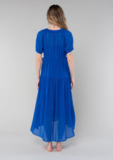 [Color: Cobalt] A back facing image of a blonde model wearing a romantic and flowy bright blue sheer chiffon bohemian maxi dress. With short sleeves, a v neckline, a high low hemline, and a drawstring waist with adjustable side ties.