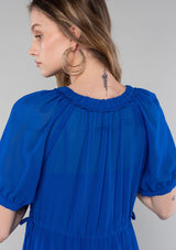[Color: Cobalt] A close up back facing image of a blonde model wearing a romantic and flowy bright blue sheer chiffon bohemian maxi dress. With short sleeves, a v neckline, a high low hemline, and a drawstring waist with adjustable side ties.