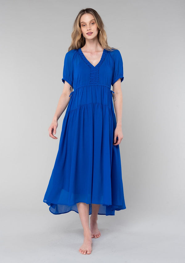 [Color: Cobalt] A front facing image of a blonde model wearing a romantic and flowy bright blue sheer chiffon bohemian maxi dress. With short sleeves, a v neckline, a high low hemline, and a drawstring waist with adjustable side ties.