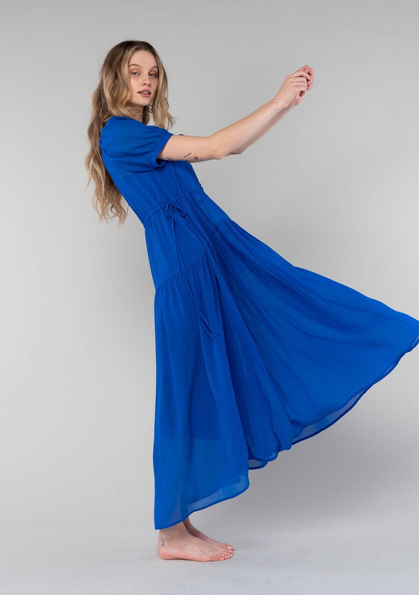 [Color: Cobalt] A full body side facing image of a blonde model wearing a romantic and flowy bright blue sheer chiffon bohemian maxi dress. With short sleeves, a v neckline, a high low hemline, and a drawstring waist with adjustable side ties.
