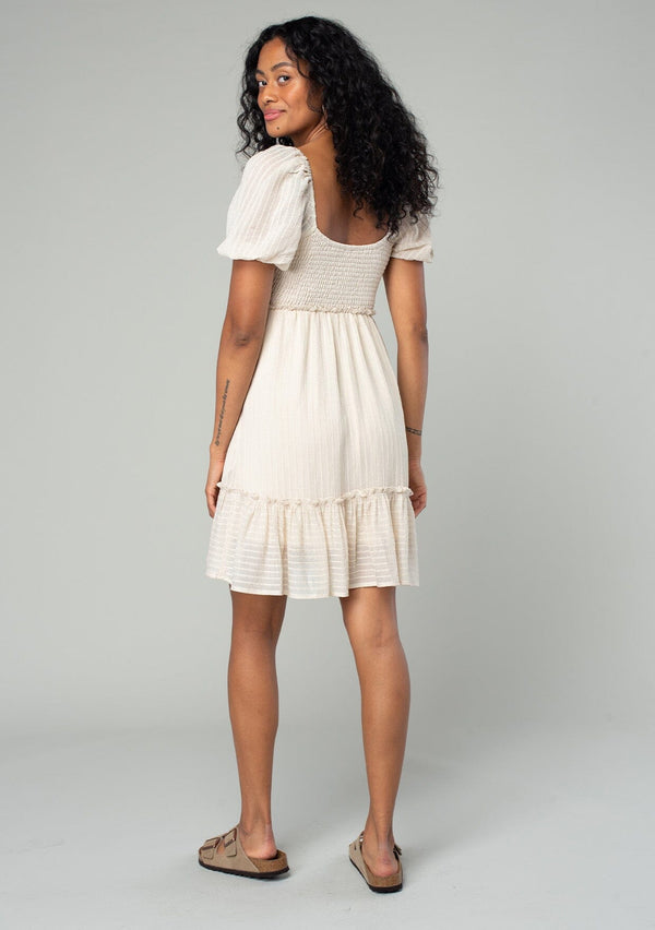 [Color: Vanilla] A back facing image of a brunette model wearing a cream colored bohemian mini dress in a textured stripe. With sheer short puff sleeves, a slim fitting smocked bodice, a flowy tiered skirt, and a square neckline. 