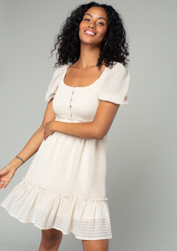 [Color: Vanilla] A front facing image of a brunette model wearing a cream colored bohemian mini dress in a textured stripe. With sheer short puff sleeves, a slim fitting smocked bodice, a flowy tiered skirt, and a square neckline. 