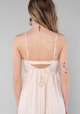 [Color: Natural] A close up back facing image of a blonde model wearing a bohemian spring maxi tank dress in a natural shadow stripe. With adjustable spaghetti straps, a round neckline, a tiered flowy skirt, a back cutout detail, and a back tie waist detail. 