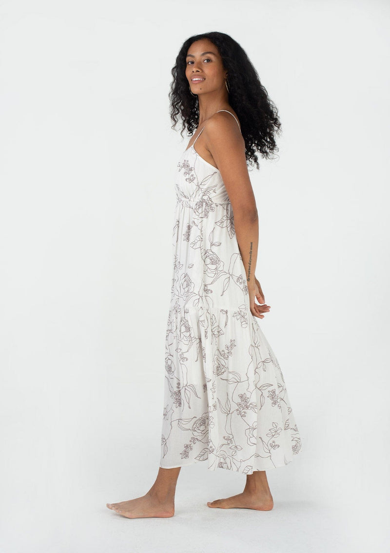 [Color: Natural/Taupe] A side facing image of a brunette model wearing a sleeveless spring maxi dress in an ivory and taupe floral print. With adjustable spaghetti straps, a scoop neckline, a flowy tiered skirt, a back cutout detail, and a drawstring waist tie at the back. 