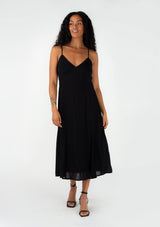 [Color: Black] A front facing image of a brunette model wearing a sexy spring slip dress in black. With adjustable spaghetti straps, a v neckline, lace trim, a half elastic waist at the back, and a flowy paneled mid length skirt. 