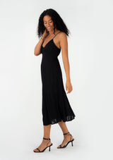 [Color: Black] A side facing image of a brunette model wearing a sexy spring slip dress in black. With adjustable spaghetti straps, a v neckline, lace trim, a half elastic waist at the back, and a flowy paneled mid length skirt. 