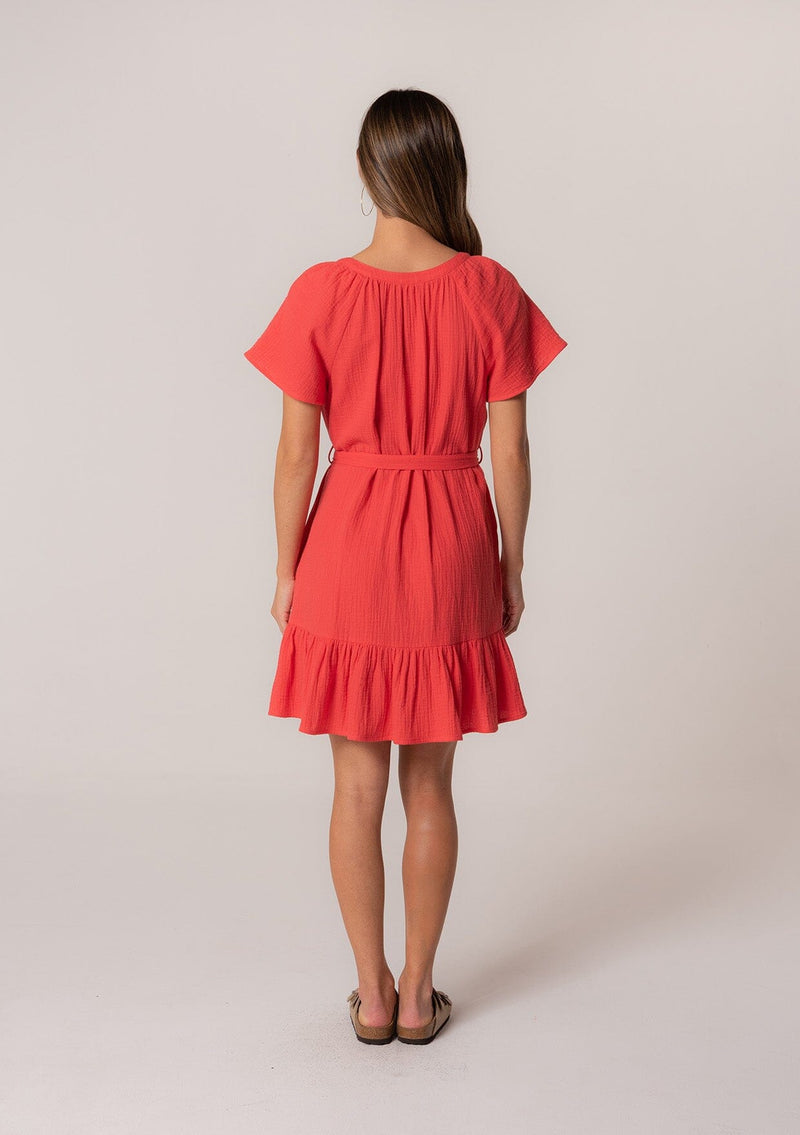 [Color: Flame] A back facing image of a brunette model wearing a spring solid red mini dress designed in cotton gauze. With short sleeves, a button front top, a v neckline, a tiered mini skirt, and a tie waist belt. 
