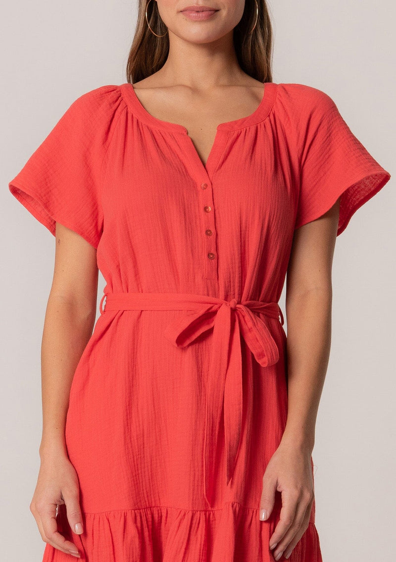 [Color: Flame] A close up front facing image of a brunette model wearing a spring solid red mini dress designed in cotton gauze. With short sleeves, a button front top, a v neckline, a tiered mini skirt, and a tie waist belt. 