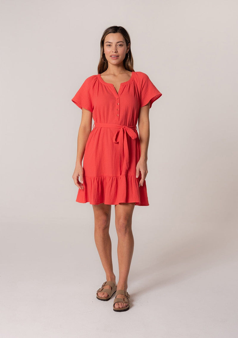 [Color: Flame] A front facing image of a brunette model wearing a spring solid red mini dress designed in cotton gauze. With short sleeves, a button front top, a v neckline, a tiered mini skirt, and a tie waist belt. 